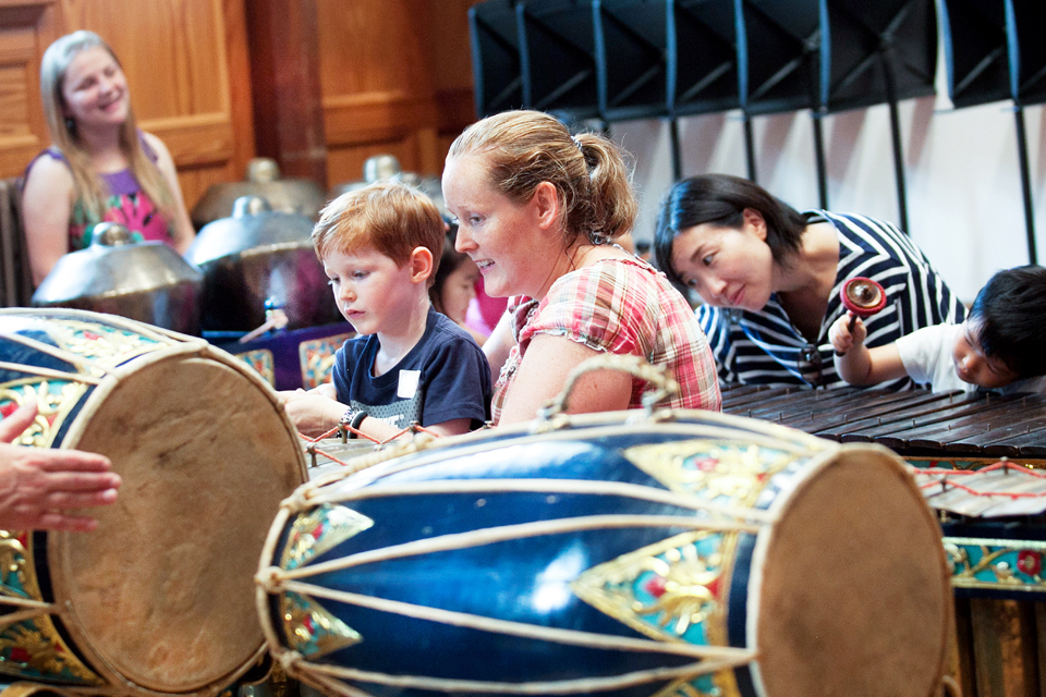 A women helping a young boy play on a xylophone, with other children and parents in the background doing the same thing.